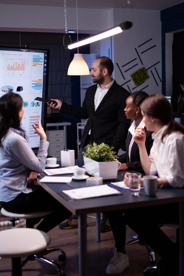 Entrepreneur man brainstorming management strategy working hard in meeting office room late at night. Diverse multi-ethnic business team looking at financial company presentation on monitor.