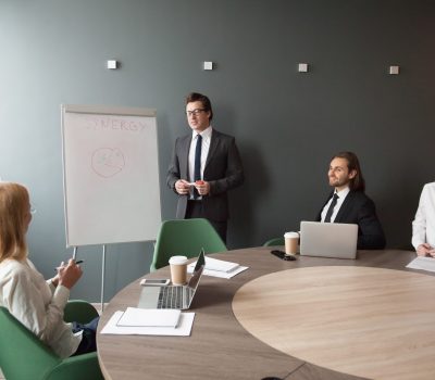 Serious middle aged businessman presenting company business goals to colleagues during meeting in boardroom, male coach training workers, explaining successful strategies on flipchart during briefing